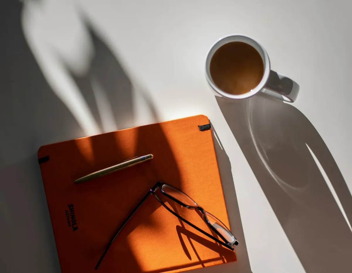Ein orangefarbenes Notizbuch mit Füller und Lesebrille, daneben eine Tasse Kaffee im Nachmittagslicht